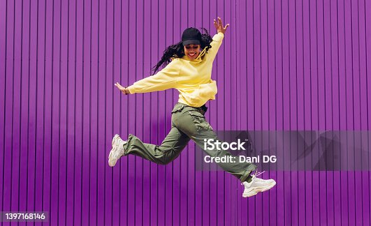 istock Jumping young woman in yellow dress on purple background. Dancer of hip hop and trap music. Concept of freedom, celebration, joy. 1397168146