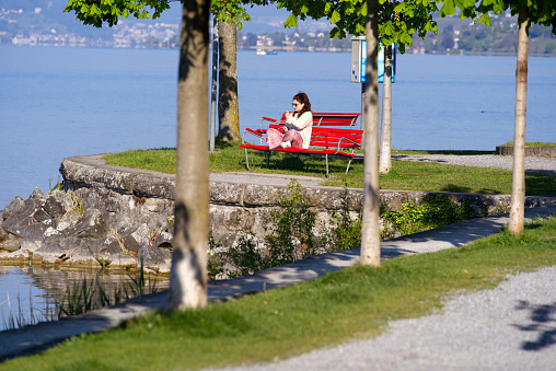 Woman with sunglasses sitting on red bench at city of Rapperswil-Jona on a sunny spring morning. Photo taken April 28th, 2022, Rapperswil, Switzerland.