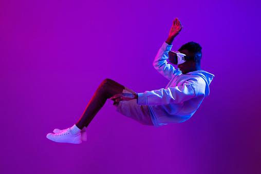 Full body side view of African man in VR goggles levitating on purple background while exploring virtual reality in studio