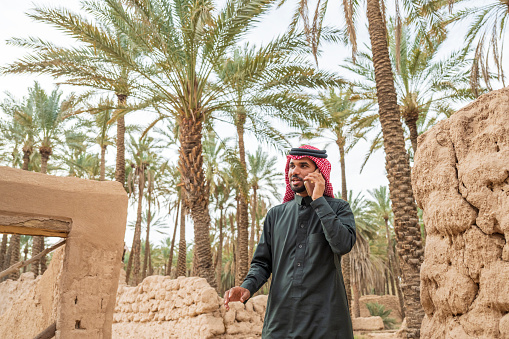 Front view of young Middle Eastern man in black dish dash, kaffiyeh, and agal talking with caller on path leading through desert oasis in Al-Ula Valley.