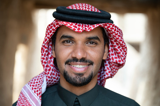 Middle Eastern man with black hair, mustache and goatee, wearing dish dash, kaffiyeh, and agal, and looking at camera with cheerful expression.