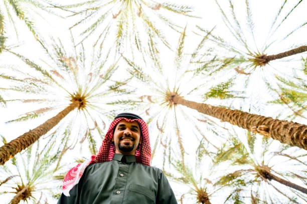 Waist-up view of bearded Middle Eastern man in traditional attire standing in sun-filled date palm grove and smiling at camera.