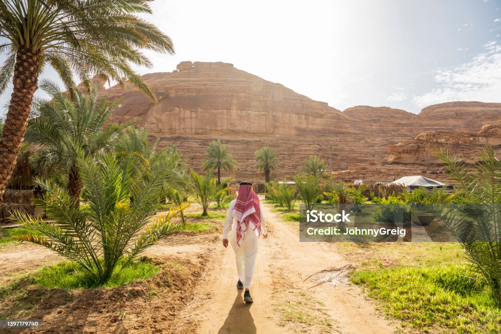 Farm worker walking to agricultural fields in Al-Ula oasis Full length rear view of Middle Eastern man in traditional clothing following dirt road to farming area beneath sandstone cliffs in Hijaz Region. Saudi Arabia Stock Photo