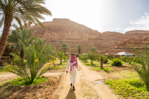 Farm worker walking to agricultural fields in Al-Ula oasis Full length rear view of Middle Eastern man in traditional clothing following dirt road to farming area beneath sandstone cliffs in Hijaz Region. arabian peninsula stock pictures, royalty-free photos & images