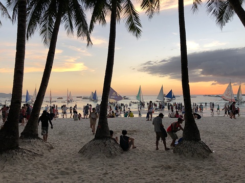 Beautiful sunset view full of tourists at the beach with palm trees, sailboats and yachts in Boracay, Philippines