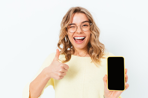 Cheerful young woman holding smartphone with blank screen and showing thumbs up