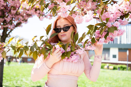 Close-up portrait of bright young woman with pink hair and sunglasses near the blossoming spring tree.