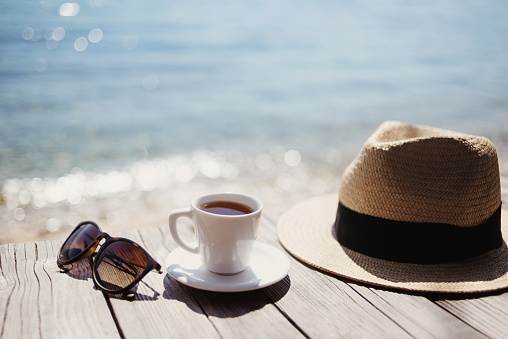 Coffee cup, sunglasses and straw hat on a table at mediterranean cafe. Travel, tourism, vacations, holidays concept