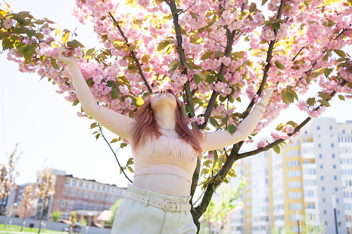 Young beautiful hipster woman near sakura blooming tree. Pink flowers, spring and youth concept. Stylish girl smiling. Blooming garden with cherry-tree