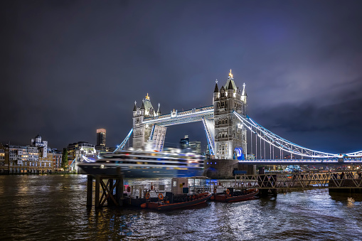 Night view of the illuminated skyline of London with a motion blurred cruise ship passing under the lifted Tower Bridge, England