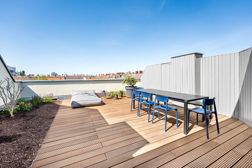 View of a rooftop deck with urban garden and view over the city skyline.  In the foreground is a dinning table with chairs. The sun is shinning and the sky is blue. There is also a rooftop garden with plants.