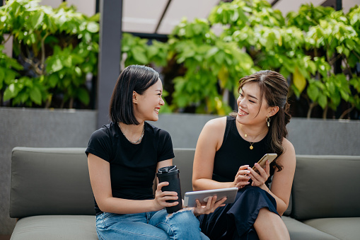 Two young Asian businesswomen using digital tablet and smartphone while having a discussion with colleagues