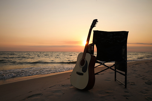 Camping chair and guitar on sandy beach near sea, space for text