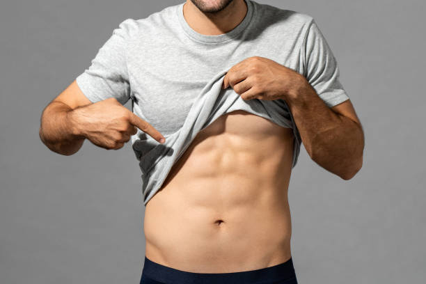 Muscular male model lifting up t-shirt to show abs while standing on gray isolated background in studio stock photo