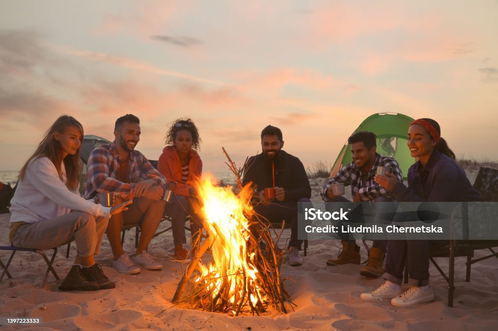 Friends sitting around bonfire on beach in evening Bonfire Stock Photo