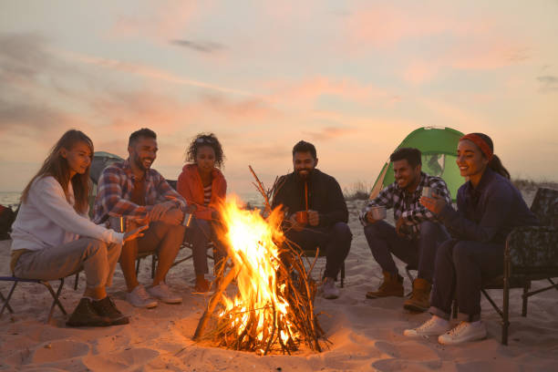 amigos sentados em torno da fogueira na praia à noite - bonfire people campfire men - fotografias e filmes do acervo