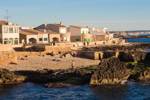 Person walking street path toward coastal landscape, carrying backpack in historic Cadaqués residential neighborhood, Mediterranean setting