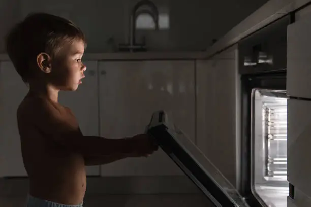 Cute curious baby boy opens the hot oven. Concept of safety and possible problems with unattended children.