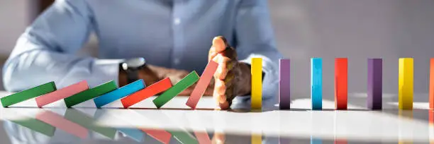 Close-up Of A Businessman Hand Stopping Colorful Dominoes From Falling On Office Desk