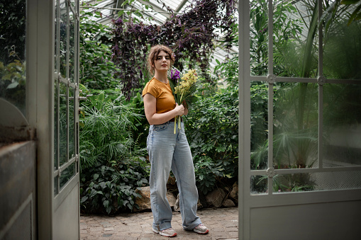 Portrait of a casually dressed female botanical worker working in a local botanical garden, holding a bouquet of flowers in her hands while standing in the middle of a botanical garden surrounded by lush green plants