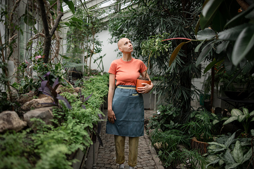 Young female botanical worker working in a local botanical garden, holding a potted plant in her hands while walking around a botanical garden surrounded by lush green plants and inspecting them