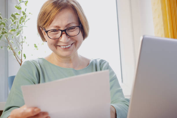 istock Happy middle aged elderly woman in eyeglasses reading paper document or postal letter from friend, sitting with laptop at home. Rejection of pleasant paperwork bank notice or post correspondence 1397236184