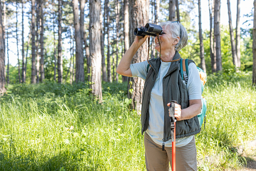 Senior woman leaning onto a hiking pole and using binoculars on a hike