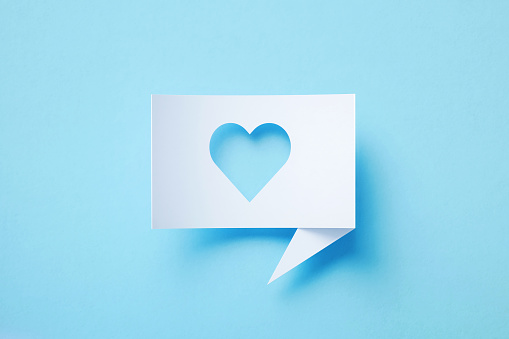 Rectangular shaped white chat bubble with cutout heart symbol sitting on blue background. Horizontal composition with copy space.