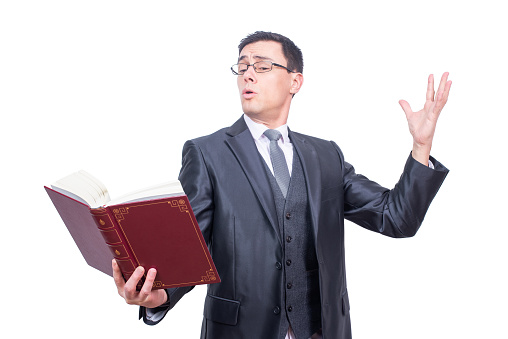 Focused male in formal suit and eyeglasses reading book aloud with raised hand while standing isolated on white background in studio