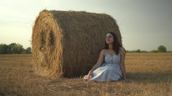 A young woman sits near a haystack in a field. A girl with glasses and a blue dress is resting in the field. 4k