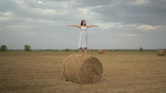 A young woman stands on a haystack in a field and spread her arms. Girl with glasses and a blue dress. 4k