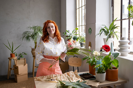 Portrait of a happy female redhead florist casually working in her flower shop, holding a bouquet of flowers in her hands, decorating it and looking at the camera with a toothy smile on her face