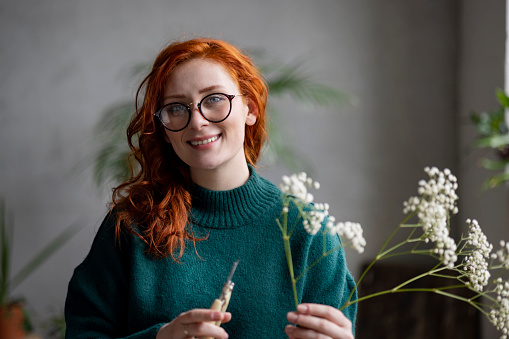 Portrait of a happy female redhead florist casually working in her flower shop, using pruning sheers to carefully cut and decorate the flowers in her hand while looking at the camera and smiling