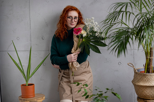 Young female redhead florist casually working in her flower shop, holding a bouquet of flowers in her hands and leaning on the wall behind her