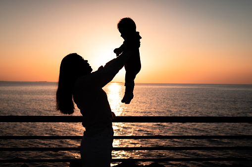 Asian woman lifting her baby boy by the seaside on the beach on a summer vacation backlit silhouette. Mother and son on the seaside spending time outdoors