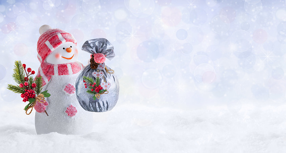 Winter or Christmas panoramic background with happy snowman dressed in pink mittens, hat and scarf in winter landscape. Christmas and New Year card or banner with a snowman holding a bag with gifts and a fir branch decorated with berries. Defocus light.