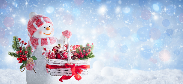 Happy snowman holding a basket with Christmas ornaments and a fir branch decorated with berries on winter snowy day. Christmas panoramic background or greeting card with snowman dressed in pink mittens, hat and scarf. Defocused light.