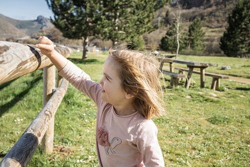 Blonde girl in beautiful nature park standing next to wooden fence playing with small insect. A family walk on Sunday afternoon, enjoying the beautiful weather.
