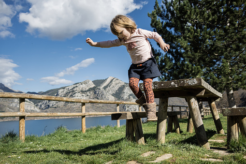 Little girl dressed in warm clothes jumping from wooden table with beautiful lake in background. Out to enjoy nature, relaxing while the beautiful scenery surrounds the place. scenery surrounds the place.