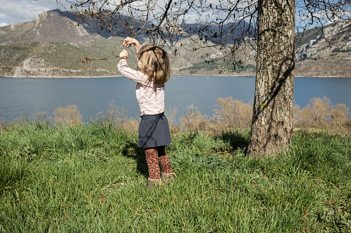 Little girl on the shore of a lake grabbing an insect from a tree branch. Girl in love with wildlife, enjoying a day in nature.