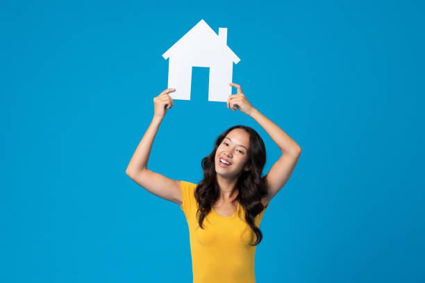 азиатская женщина поднимает модель дома, стоя на синем фоне в светлой студии - allegory painting human settlement built structure house стоковые фото и изображения