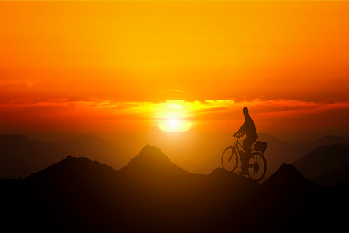 Leadership and goals. A man standing on top of a mountain on a bicycle watching the sunset. Conceptual image composite.