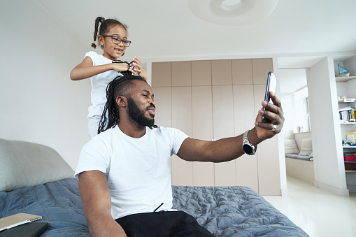 Pleased adult man looking at phone while daughter playing with his hair