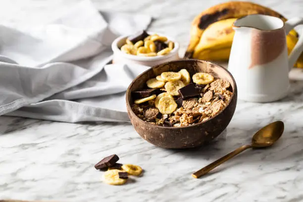 Muesli made from mix of unprocessed whole grains, chia, quinoa, nuts, seeds, with banana and chocolate in coconut vowl on the marble table . Heathy breakfast concept.