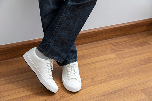 Legs of a young man in blue jeans and white sneakers on wood floor.
