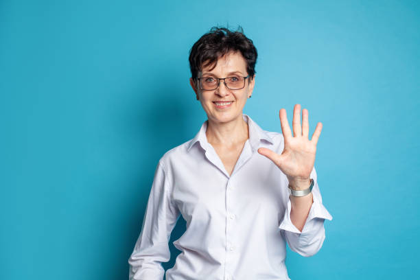 Portrait of smiling middle adged woman in glasses showing five fingers against blue studio wall background Portrait of smiling middle adged woman in glasses showing five fingers against blue studio wall background i 5 stock pictures, royalty-free photos & images