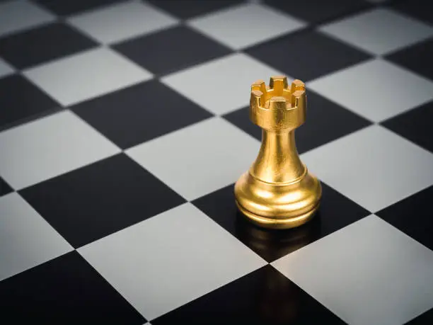 Photo of The golden rook chess piece standing alone on chess board background. Rook or the castle, the tower symbolize a protectorate of the city.