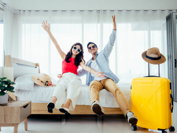 여행 준비, 행복한 휴가, 체크인. 여름 방학 개념. 기쁨으로 손을 들어 올리는 아시아 커플, 남자와 여자는 여행 중에 노란색 여행 가방이있는 하얀 침대에서 행복하게 선글라스 미소를 짓습니다 - hotel room 뉴스 사진 이미지