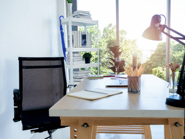 istock Modern workplace with empty black chair, pencil, notebook table lamp and laptop computer on wood desk near the book shelf and glass window, office, white . 1397293639