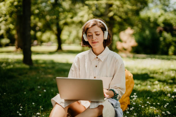 A loving and cute young adult student, a brunet, sitting in the park with a white headphones on her head, listening to a music while typing on her laptop. She's urban, casually dressed, having an yellow ocher back pack next to her, and being focused while enjoying the sun rays